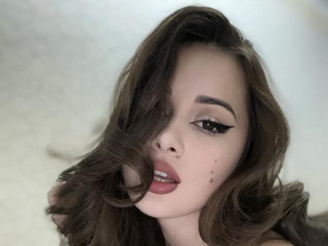JayerSey has wavy brown hair covering one eye, dark eyeliner, and red lips. She has small moles on her left cheek. Her expression is slightly pouty.