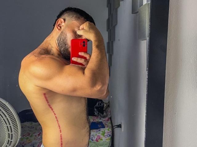 TeamLatin, shirtless, muscular, holding red phone, covering face, tattooed side, scar down back, standing in front of mirror, short black hair, beard, in bedroom, visible fan.