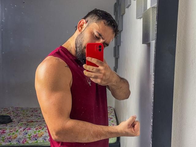 TeamLatin with a muscular build, dark curly hair, and a red tank top, holding a red phone, taking a mirror selfie, showing a flexed arm.