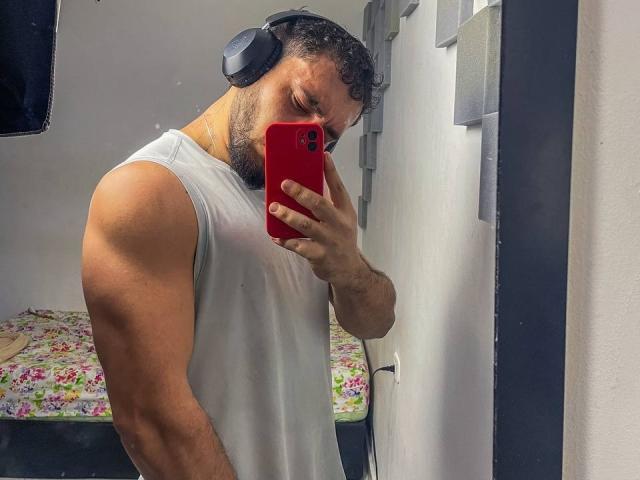 TeamLatin with a muscular build, wearing a white sleeveless shirt, black headphones, and a red phone, taking a mirror selfie. His arm is flexed, showing a tattoo.