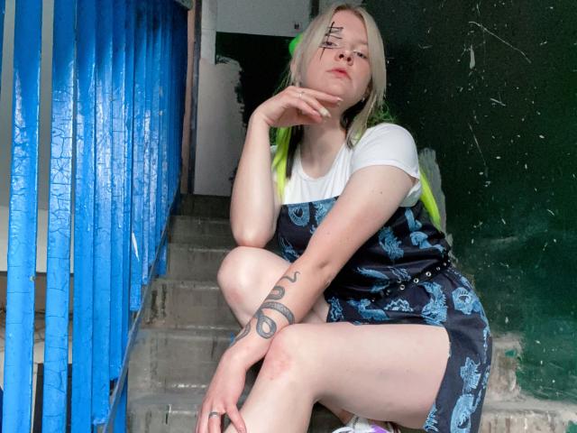HettieNell, avec ses cheveux blonds-vert, son t-shirt blanc, sa robe fleurie bleue, son bras tatoué, assise sur des marches en béton, la main sur le menton, une rambarde bleue à gauche, un mur vert en arrière-plan..