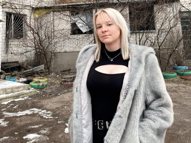HettieNell with blonde hair, wearing a gray fur coat over a black cutout dress, showing her tits, standing in a dirty, snowy outdoor area.