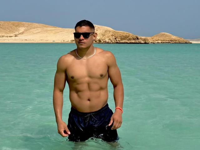 LoganVelasco standing in clear water, shirtless, with a toned, muscular chest and abs, wearing black shorts and sunglasses, a red bracelet on his right wrist.