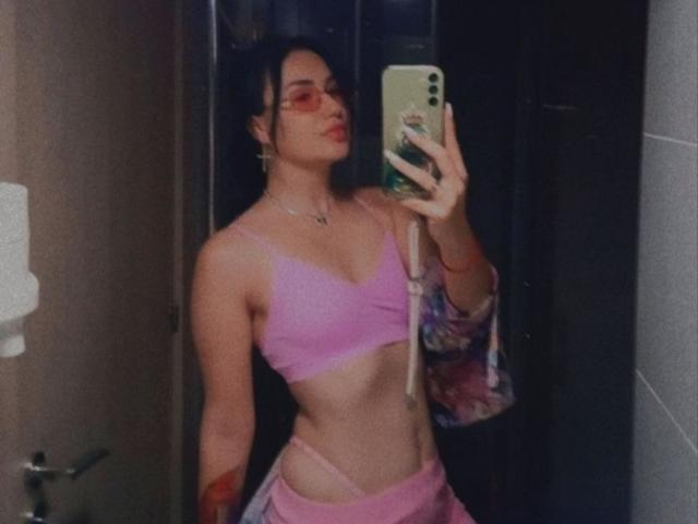 VioletChriss with pink sports bra, showing her tits, pink pants, and tattoo, taking a mirror selfie with a phone, wearing glasses, and a floral jacket.