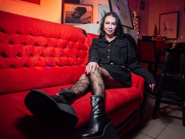 VioletChriss sits on a red couch, wearing a black dress, patterned tights, and black boots, with her legs crossed, showing her black heels and a subtle smile.