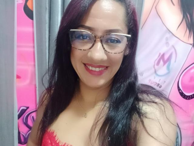 SamanthaCorine with long black hair, glasses, red lipstick, and a red lace bra, smiling at the camera, showing off her tits. She's wearing a gold necklace.