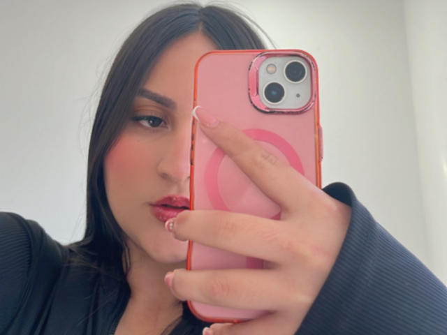 MiaCooperr mit dunklem Haar, einer rosa Handyhülle, einem roten Ring, einem schwarzen Oberteil, voller Lippen, die ein Handy hält und ein Selfie macht, vor weißem Hintergrund. Brust nicht sichtbar, Handy im Vordergrund..