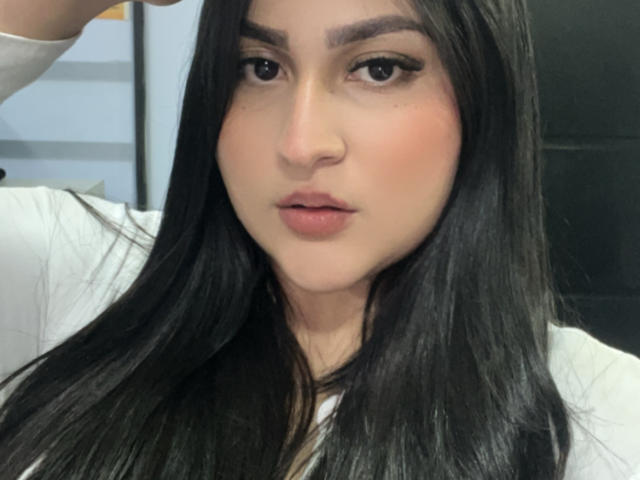MiaCooperr has long black hair, dark eyebrows, and pink lips. She's wearing a white shirt, looking directly at the camera with a slightly pouty expression.