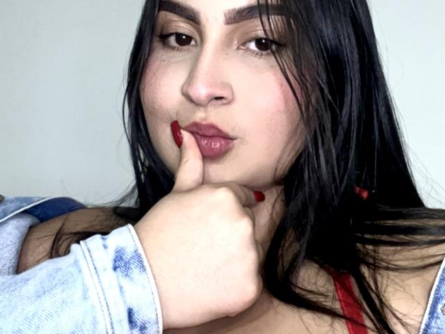 MiaCooperr, com cabelo escuro, unhas vermelhas e um dedo perto de seus lábios rosados e brilhantes, vestindo uma camisa azul e branca e uma blusa vermelha. Seus seios estão ligeiramente visíveis..