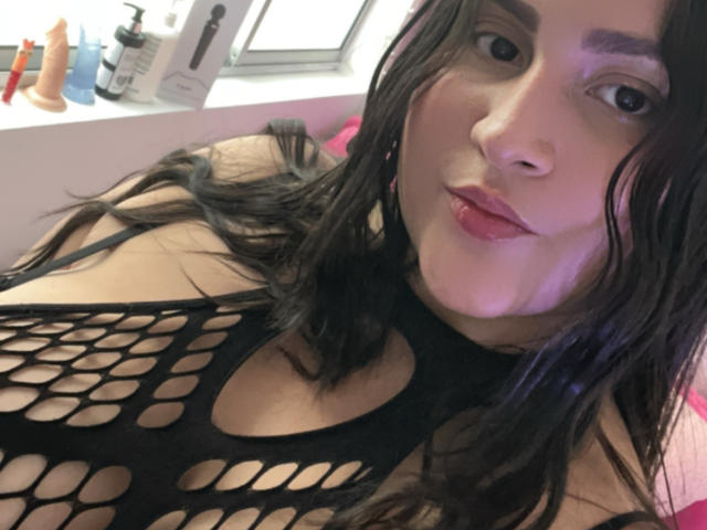 MiaCooperr draagt een zwarte fishnet top, waardoor haar borsten zichtbaar zijn. Ze heeft nat donker haar, roze lippenstift en een speelse uitdrukking. Ze zit in een kamer met een dildo en een fles lotion op een vensterbank..
