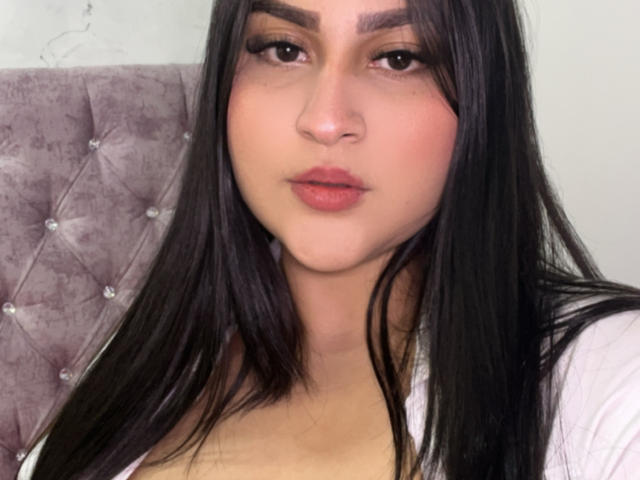 MiaCooperr, com cabelos pretos longos, olhos castanhos e lábios rosados, vestindo uma blusa branca, mostrando levemente seus seios, sentada em um encosto cinza acolchoado..