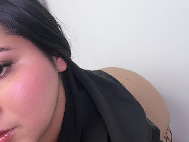 Het gezicht van MiaCooperr, met roze blush, zwart haar en een gedeeltelijk zichtbaar zwart topje, waarbij haar blote billen te zien zijn. Haar oog is lichtjes open en haar lippen zijn een beetje open..