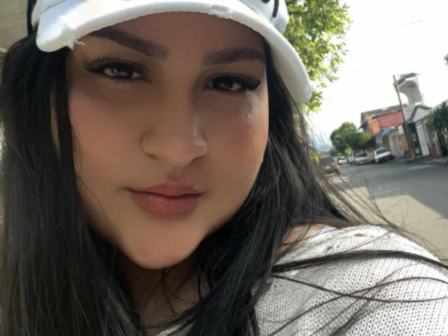 MiaCooperr hat ein süßes Gesicht mit langen, schwarzen Haaren, dunklen Augenbrauen und vollen Lippen. Sie trägt eine weiße Mütze und ein graues Hemd, mit einem unauffälligen Nasenpiercing..