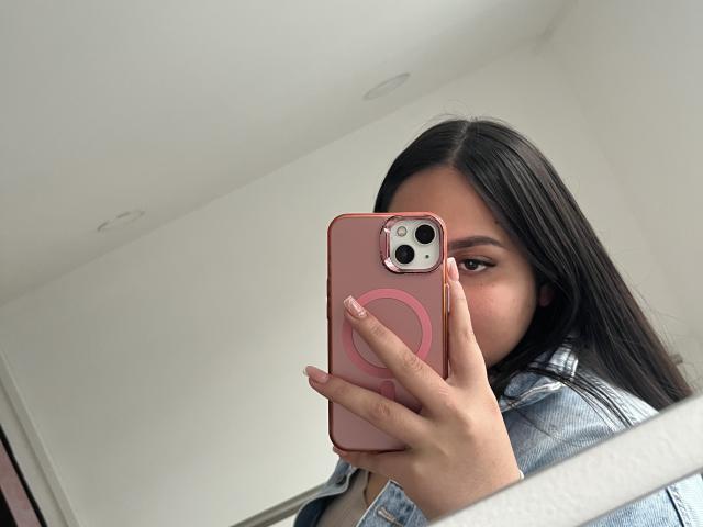 MiaCooperr macht ein Selfie mit einem rosa Handy und zeigt ihr Gesicht und einen Teil ihrer Brüste. Sie hat langes, schwarzes Haar, braune Augen und trägt eine Jeansjacke..