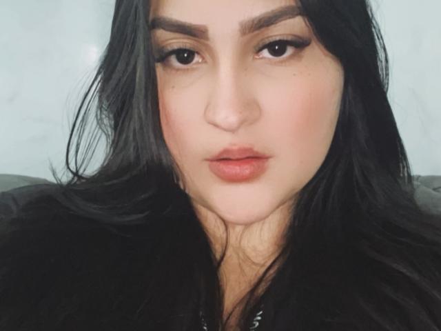 MiaCooperr heeft zwart haar, bruine wenkbrauwen en volle, roze lippen. Ze staart voor zich uit met lichtjes open, huidskleurige lippen. Haar ogen zijn donker en ze draagt subtiele make-up..