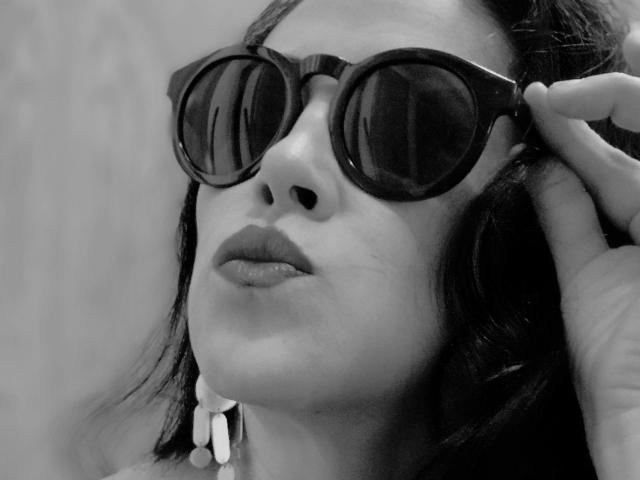 Claudias wearing dark, round sunglasses with her hand adjusting them. She has dark hair, glossy lips, and a white earring. Black-and-white image, focusing on her face.