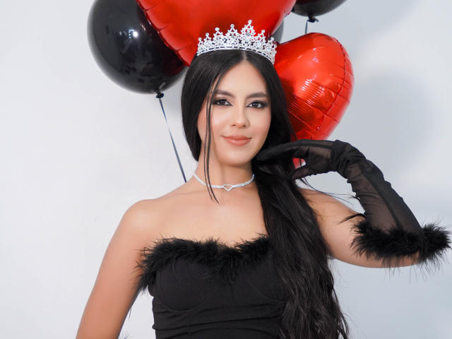 EimmyPark with long black hair, wearing a silver tiara, black strapless dress with fur, black gloves, and choker, holding red and black balloons, smiling.