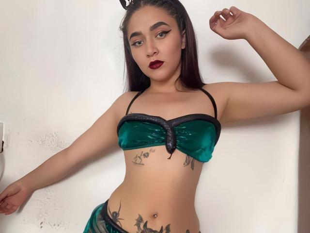 SophieHaily with dark hair, green velvet bra with black trim, tattoos on stomach and hip, arms raised, red lipstick, looking at camera, standing against white wall.