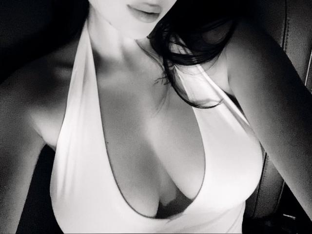 Os seios grandes e nus de Olieri, em um top branco de alças finas com decote profundo, cabelo preto cobrindo parcialmente o rosto, em uma foto em preto e branco, com alto contraste..