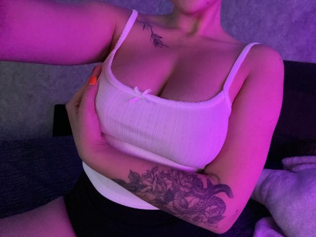 Olieri's white ribbed tank top with a small bow, covering her big tits, and black shorts. She has tattoos on her chest and arm, under pink and purple lighting.