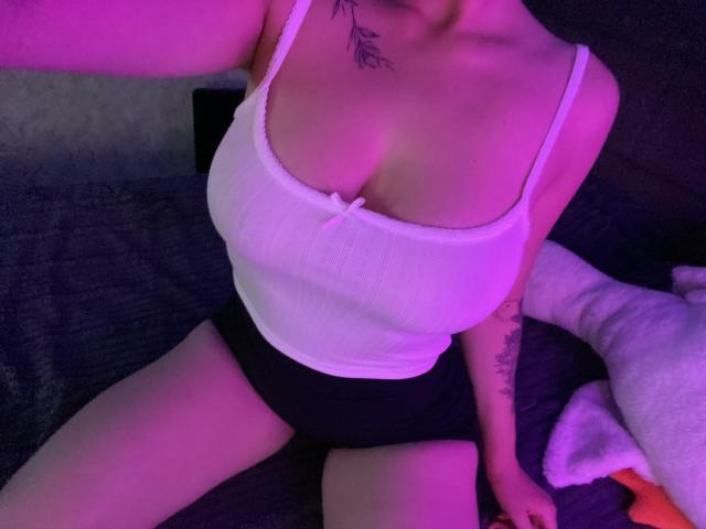 Olieri with large tits in a white ribbed tank top and black shorts, sitting with legs apart, purple lighting, tattoos on neck and arm, on a bed.