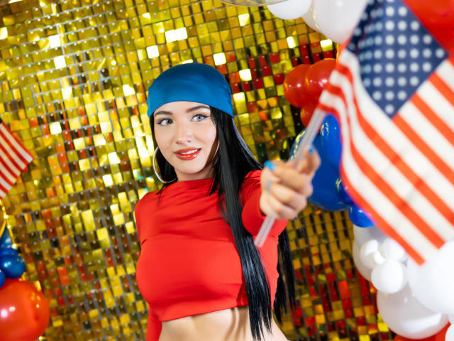 LillyTaylor with long black hair, blue headband, red crop top, and red lipstick, holding an American flag, smiling, standing in front of gold sequins and red/white/blue balloons.