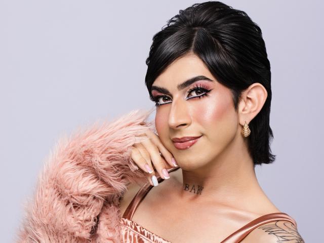 MayaPeitthon with black hair, pink eyelids, gold earrings, pink fluffy sleeve, tattoo on neck, tan skin, and pink lipstick, smiling at camera.