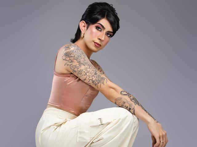 MayaPeitthon with short black hair, pink tank top, white pants, tattooed arms, and pink eyeshadow, squatting, looking at camera, showing off her tits and ass.