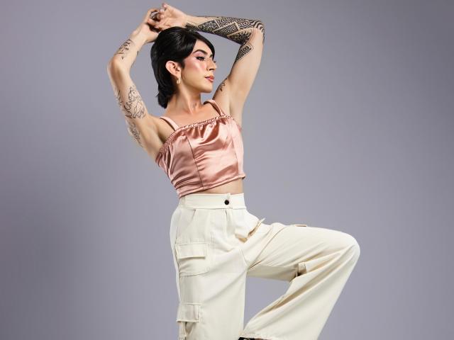 MayaPeitthon with short black hair, tattoos on arms, pink satin top, white high-waisted pants, arms raised, one leg bent, looking to the side, confident, stylish.