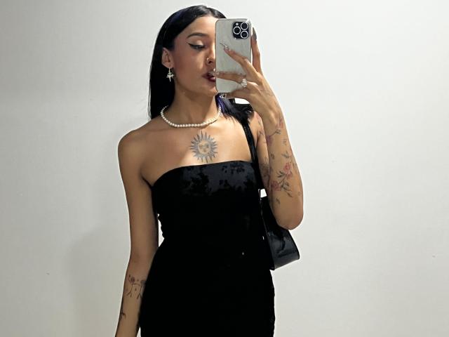 SalomeMille with long black hair, wearing a black strapless dress, pearl necklace, and earrings, holding a phone to her face, showing tattoos on chest and arm.