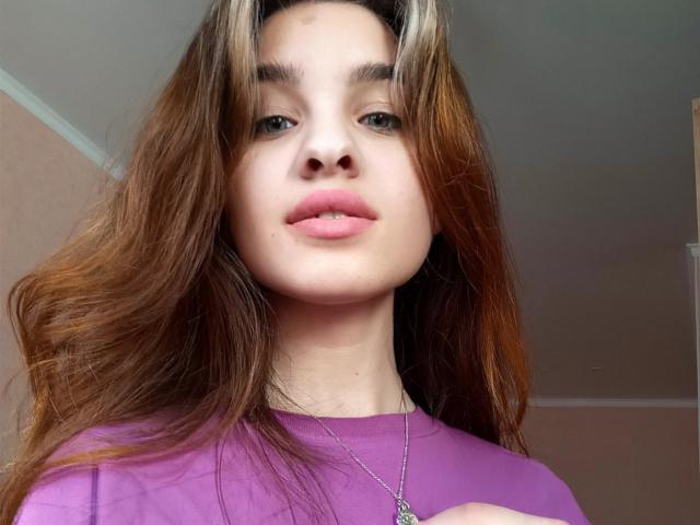 Ssimona has long brown hair, pink lips, and wears a purple shirt with a silver necklace. Her face is centered, looking slightly down at the camera.