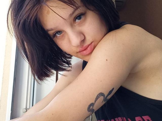 Ssimona with short brown hair, blue eyes, and a tattoo on her arm, leaning by a window, showing her bare shoulder and pouty lips, wearing a black top.