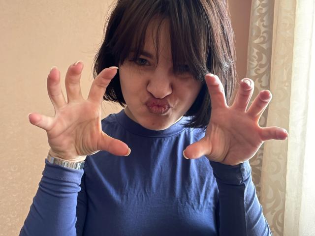 Ssimona with short brown hair, pouty lips, and blue long-sleeve shirt, hands up, fingers spread, wearing a wristwatch, looking cute and slightly goofy.