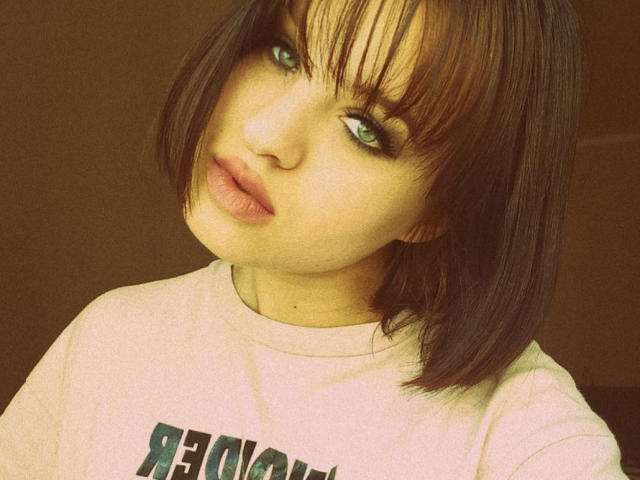 Ssimona has a cute bob with brown hair, green eyes, and pouty lips, wearing a white tee. Her tits are small and her ass is not visible.