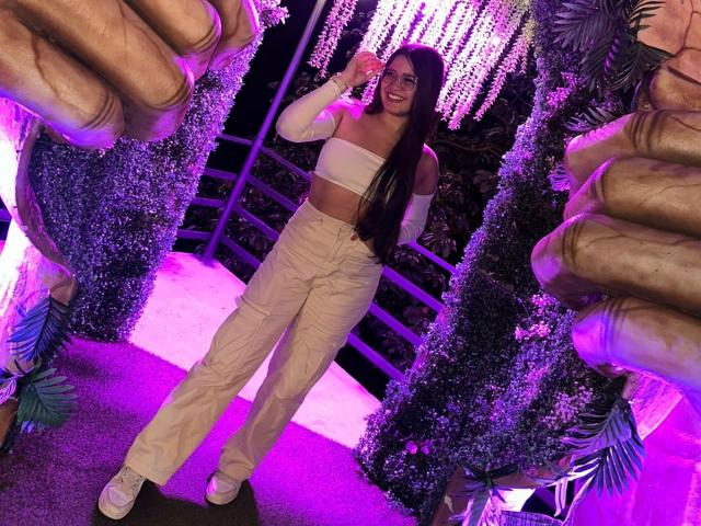 CamilaSofiia with long dark hair, white strapless top, and beige pants, smiling, holding pink drink, surrounded by giant hands, purple lighting, white sneakers, and leaf decorations.