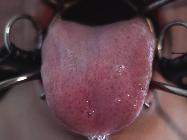 AkiraNishimura69's wet, pink tongue is being held by metal rings, showing saliva and small red specks. The image is a tight, detailed shot focusing on the tongue.