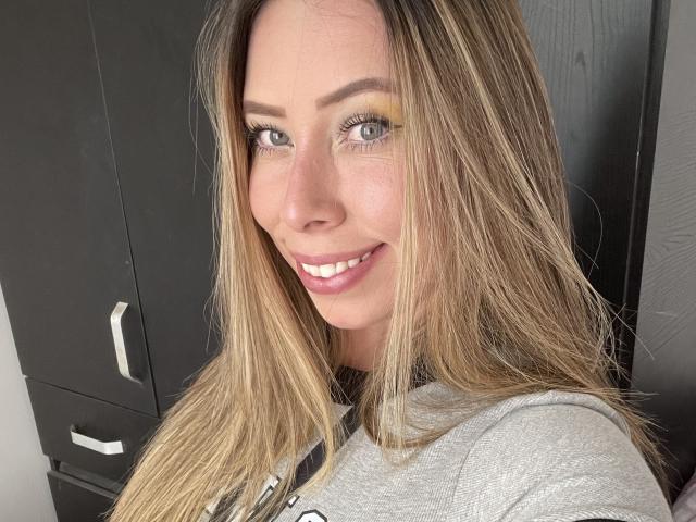 JennyLawry with blonde hair, green eyes, and yellow eyeshadow, smiling at the camera. She's wearing a gray shirt, and her tits are partially visible. Watermark in bottom right.