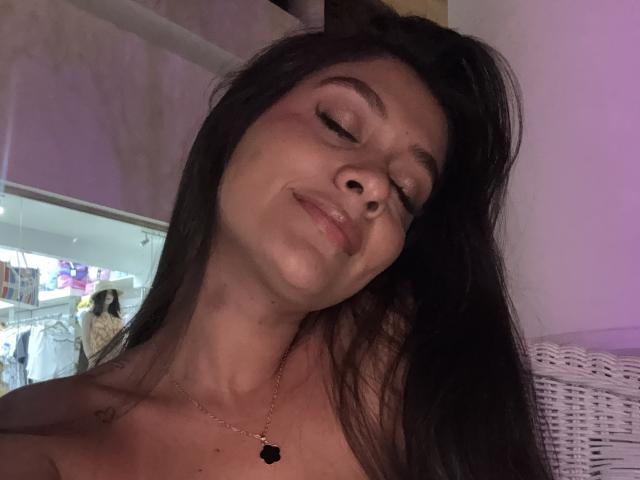 ThaliaAres has long black hair, slightly squinted eyes, and a small smile with a slight tilt to her head. She's wearing a necklace with a black pendant.
