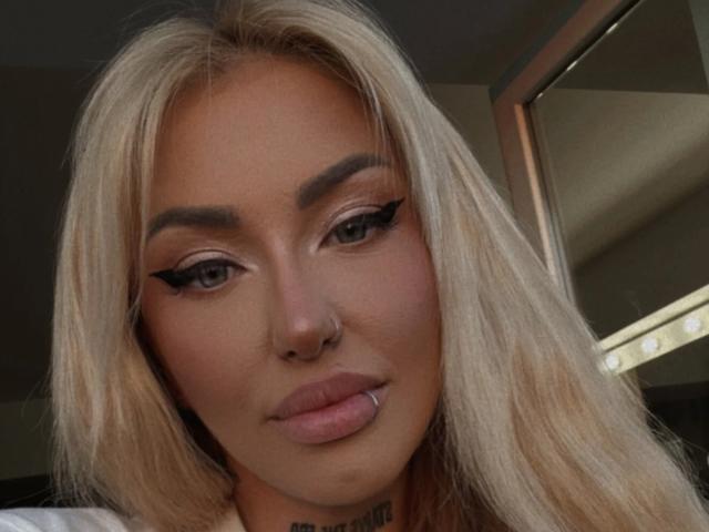 SinnersMuse has blonde hair, winged eyeliner, and a nose ring. She's sucking her pink lips with a neutral expression, showing a tattoo under her chin.