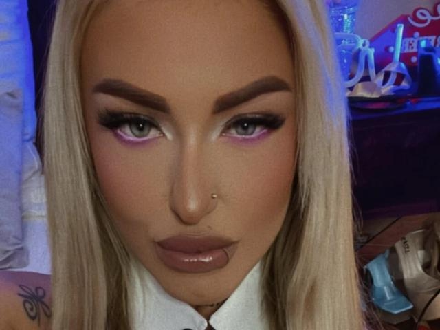 SinnersMuse has blonde hair, green eyes with pink eyeliner, thick eyebrows, a nose piercing, and glossy brown lips. She's staring directly at the camera with a serious expression.