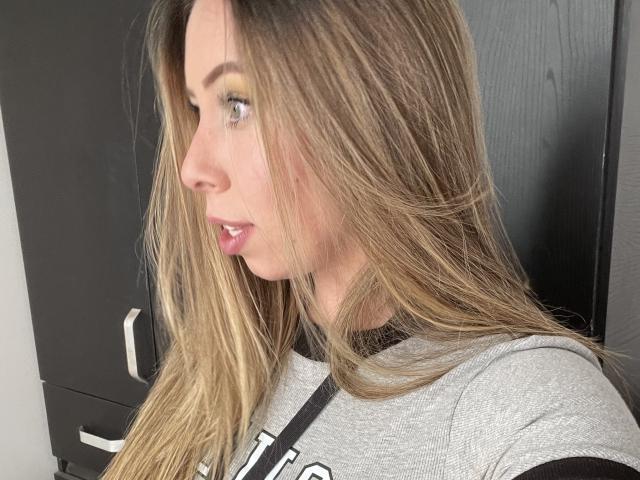 JennyLawry, con cabello rubio largo y liso, vestida con una camisa gris, vista de perfil que muestra su nariz, labios y ojos verdes, mirando hacia la izquierda..