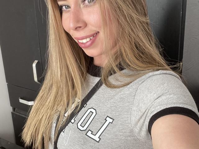 JennyLawry with long blonde hair, smiling, wearing a grey sports shirt with black trim and white lettering, showing her tits, standing in front of dark furniture.