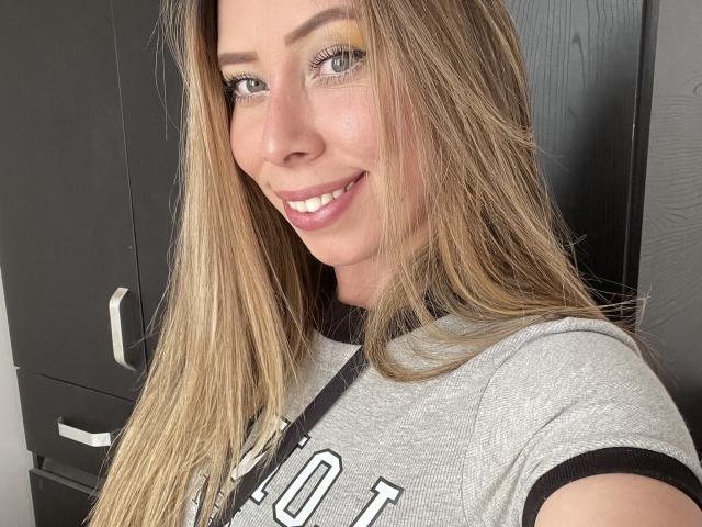 JennyLawry with blonde hair, green eyes, and a smile, wearing a gray shirt, showing her tits, standing in front of a black cabinet.