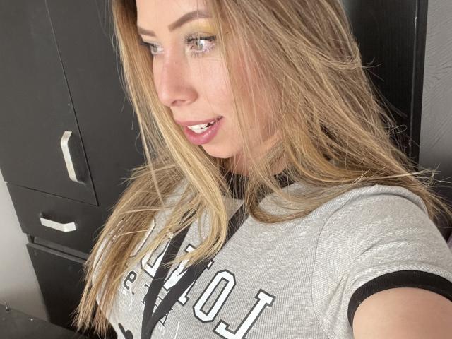 JennyLawry, com cabelo loiro, vestindo uma camisa cinza, mostrando seus pequenos seios, lábios rosados levemente entreabertos e olhos azuis, tirando uma selfie..