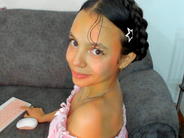 DanesaCronin with braided hair, pink off-shoulder top, holding pink keyboard and white mouse, smiling, brown eyes, nose dot, silver necklace, grey couch, small white hairclip.