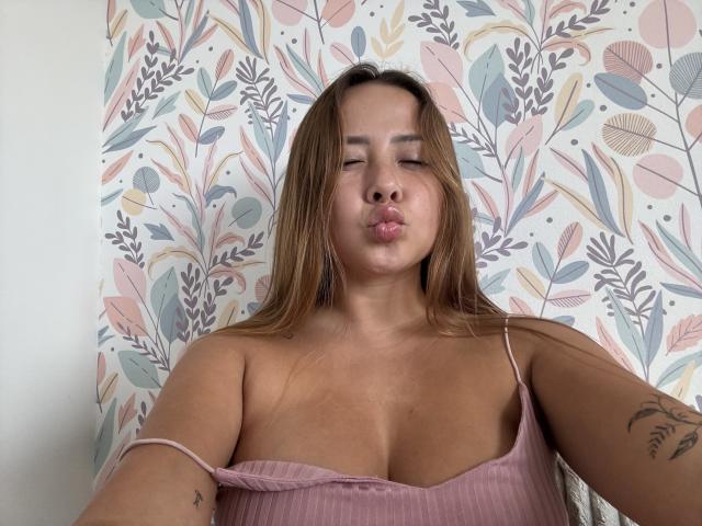 NatSorian, avec ses lèvres charnues, ses cheveux bruns et son haut rose côtelé, dévoilant sa poitrine et ses tatouages, les bras légèrement levés, les yeux marrons, une expression détendue, debout devant un papier peint floral..