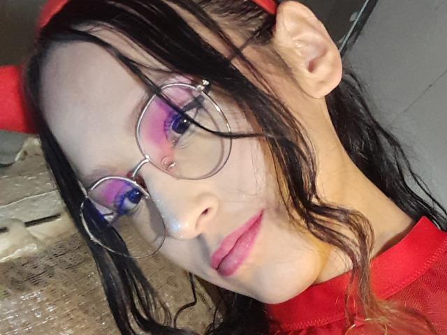 WorthyOfBdsm with glasses, pink eyeshadow, red headband, and red shirt, has wet black hair, pink lips, and a slight smile, in a selfie with a metal floor.