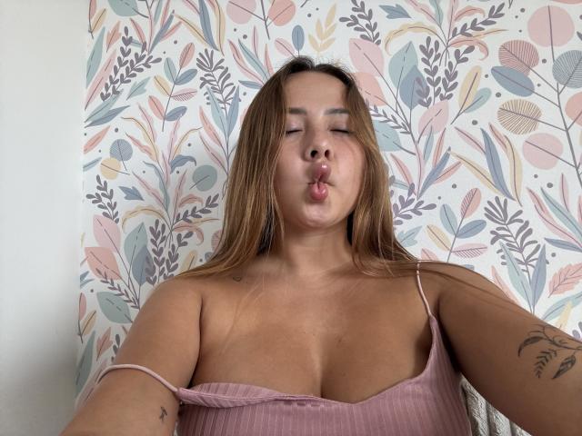 NatSorian, avec des lèvres boudeuses, des cheveux bruns, un haut rose côtelé qui dévoile sa poitrine, des tatouages sur le bras, sur un fond de papier peint floral. C'est une jeune femme ronde, mignonne, qui fait une grimace amusante..