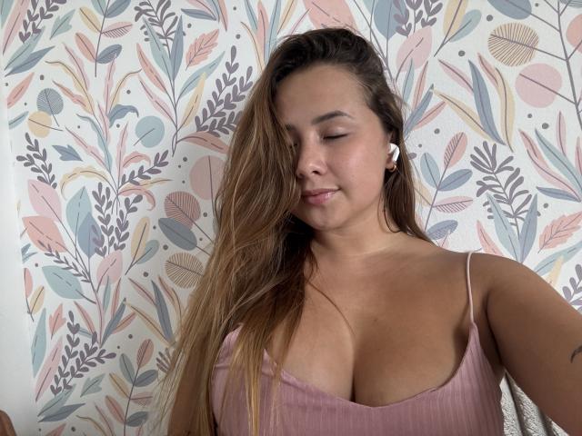 NatSorian con lunghi capelli castani, una maglietta a coste rosa che mette in risalto il suo seno prosperoso, indossa auricolari bianchi, ha gli occhi chiusi e sorride dolcemente..