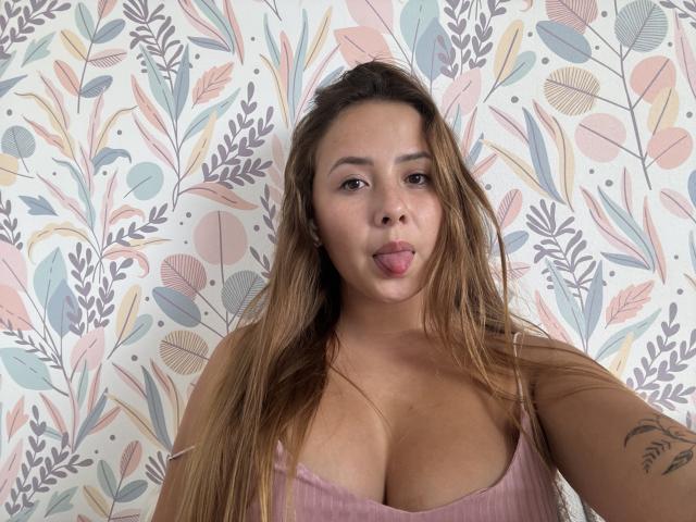 NatSorian has long brown hair, a tattoo on her arm, and is sticking out her tongue. She's wearing a pink ribbed top, showing her boobs.